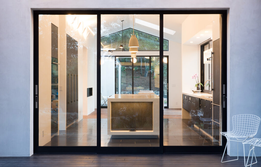 In a modern black and white kitchen with vaulted ceilings, a black-framed multi-slide glass door complimented by a large triangular window on top looks into a backyard full of trees.