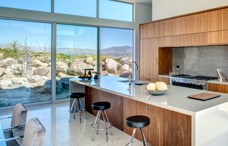 A multi-slide door peers into desert landscaping in a modern kitchen with a white waterfall island and light brown cabinets.