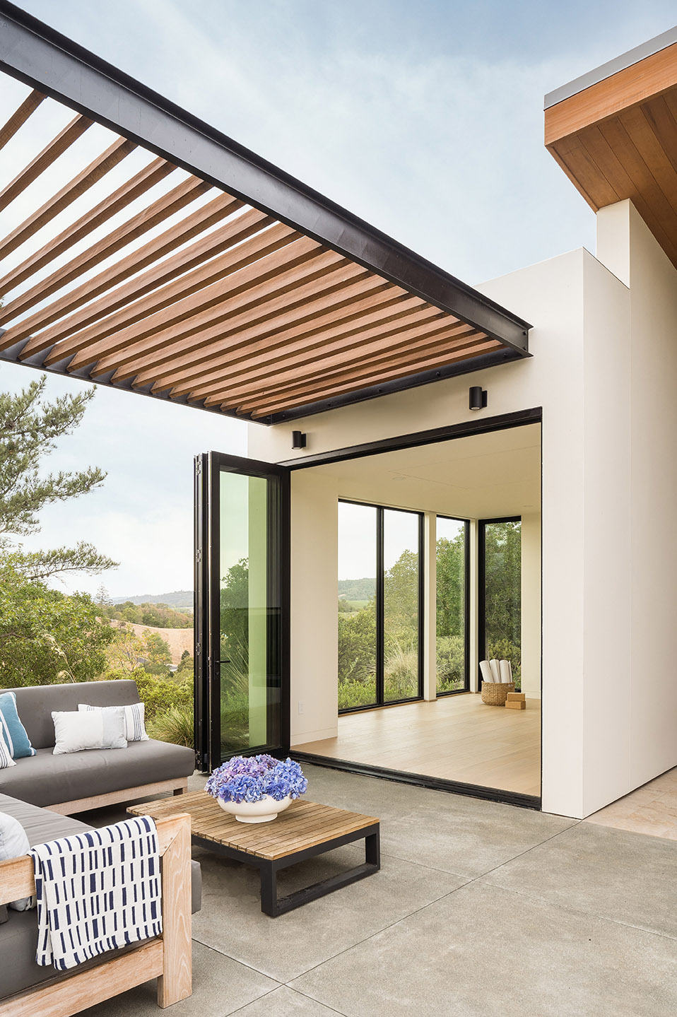 A modern, metal wrapped pergola provides shade for the outdoor seating area in front of an open bi-fold door.